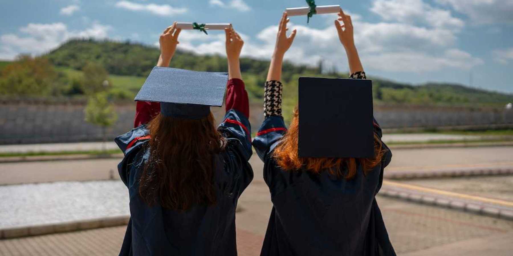 Two graduates with backs to camera holding their rolled-up diplomas above their heads, against the sky