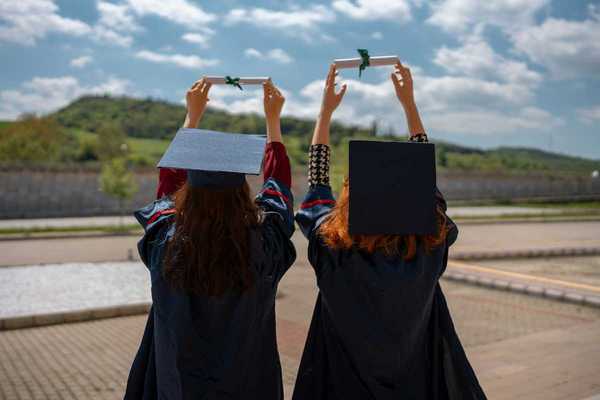 Two graduates with backs to camera holding their rolled-up diplomas above their heads, against the sky