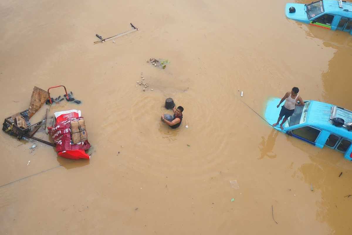 Two men attempting to salvage items inundated by floodwaters amidst submerged vehicles.