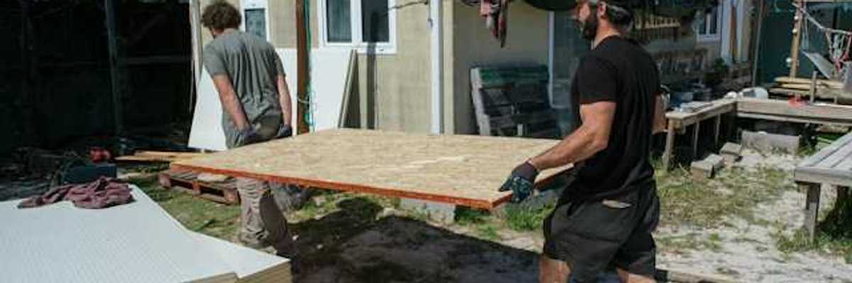 Two men carrying a plywood sheet past a manufactured home