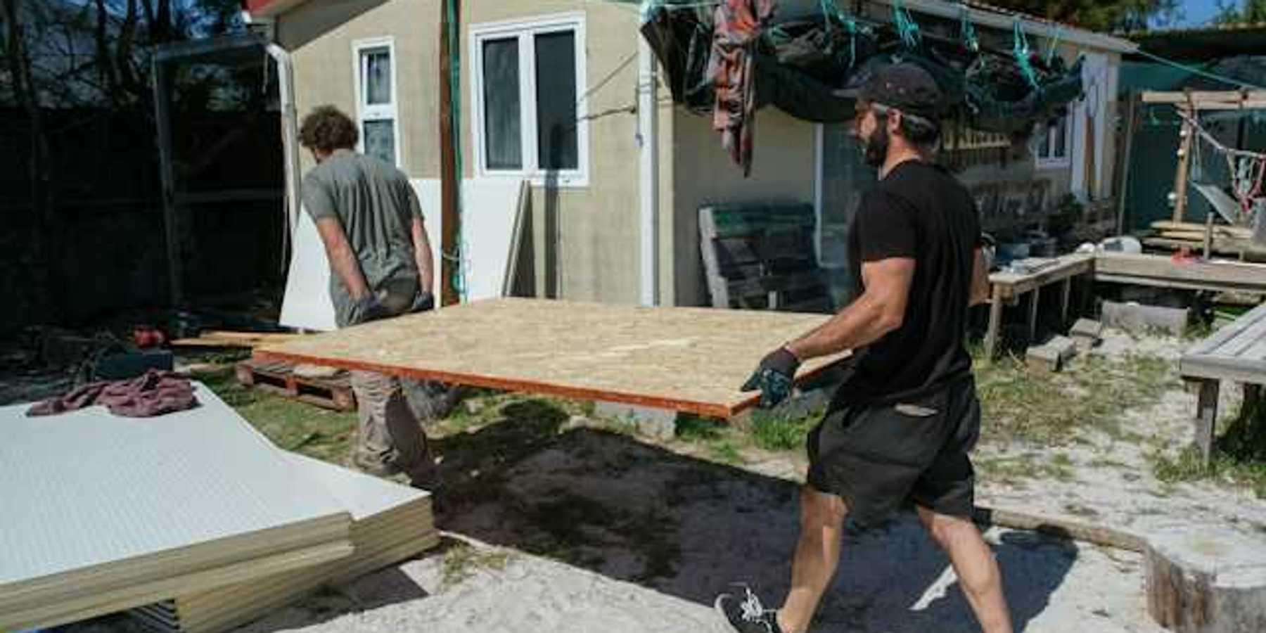 Two men carrying a plywood sheet past a manufactured home
