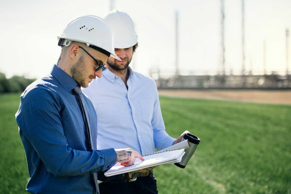 Two men conferring over a notebook in front of an oil refinery
