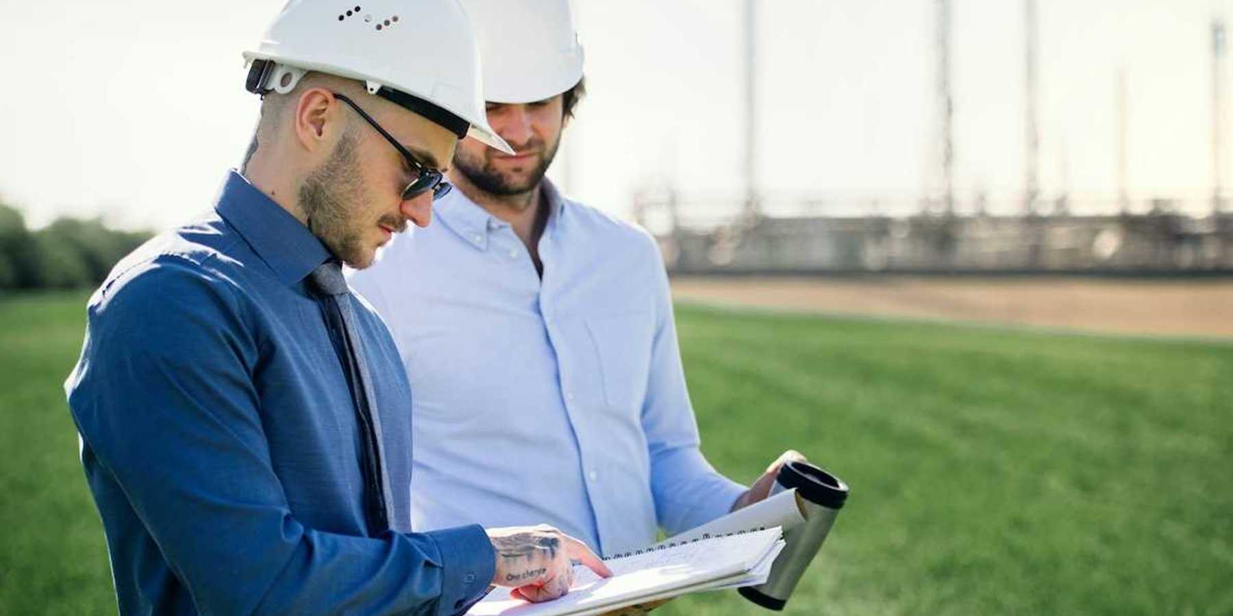 Two men conferring over a notebook in front of an oil refinery
