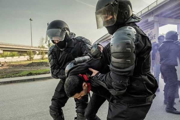 Two men dressed in protective gear - pads and helmets with face shields - while either detaining or helping a third man