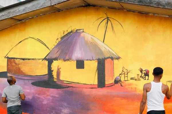 Two men painting a mural on a concrete wall of an African hut and village scene