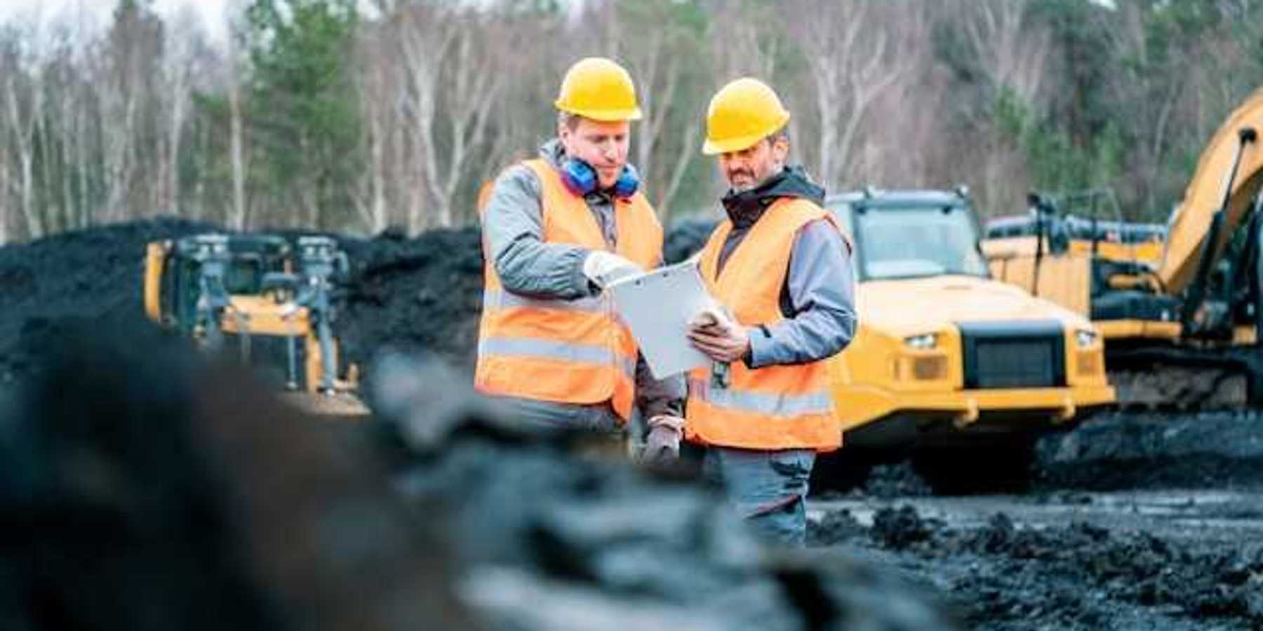 Two men wearing orange safety vests standing in front of piles of coal