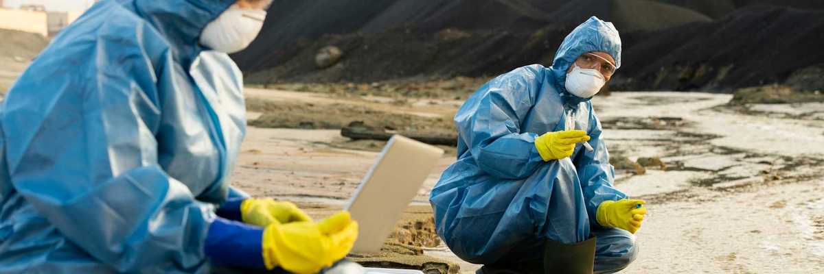 Two people clad in blue haz-mat suits sampling water running adjacent to a coal waste pile