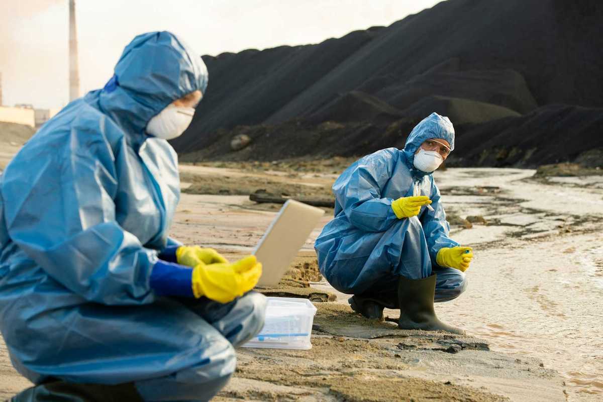 Two people clad in blue haz-mat suits sampling water running adjacent to a coal waste pile