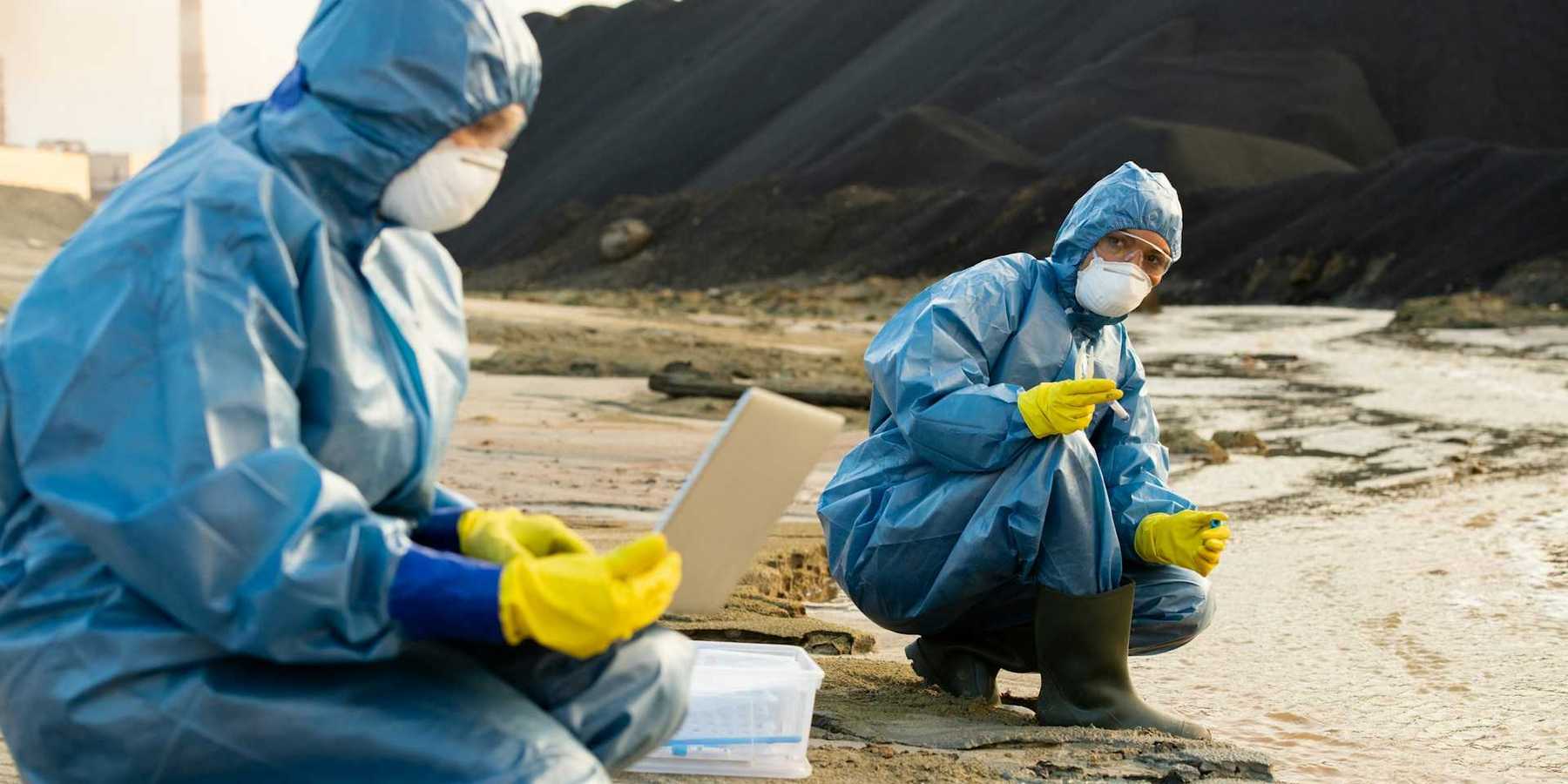 Two people clad in blue haz-mat suits sampling water running adjacent to a coal waste pile