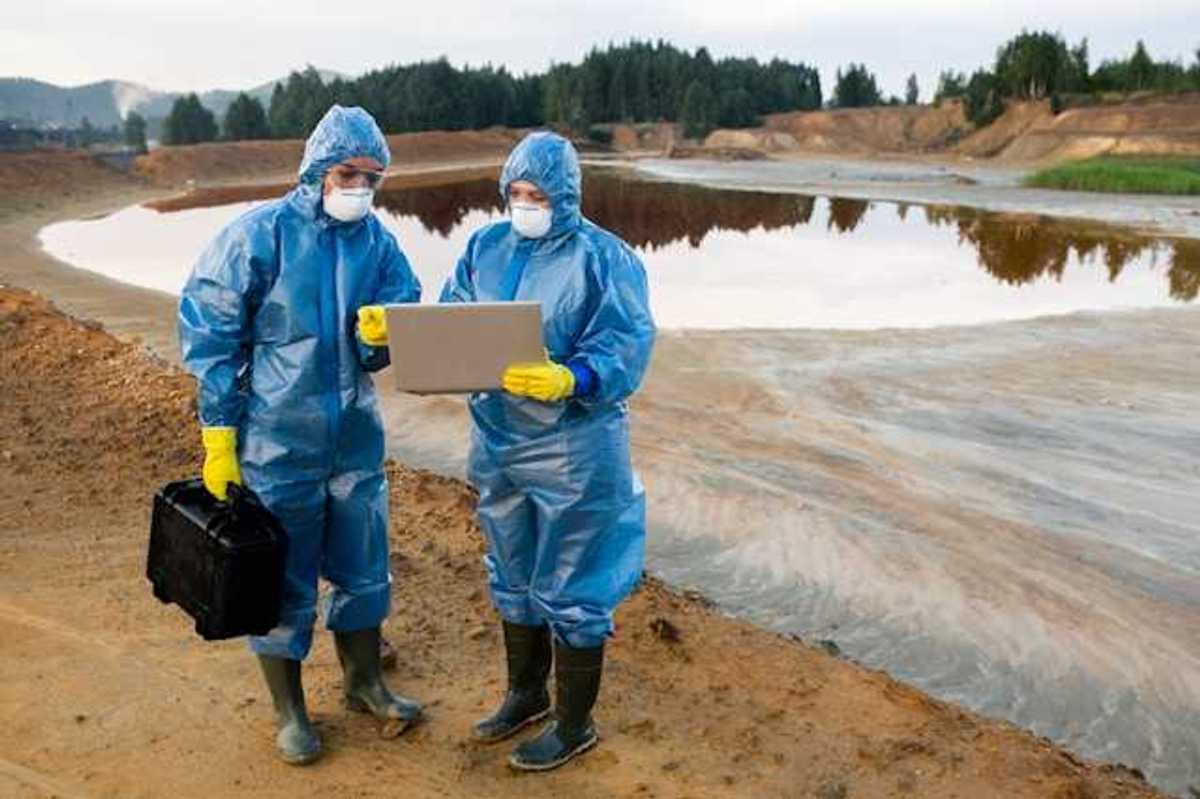 Two people wearing protective gear looking at a laptop in front of a mining waste pond