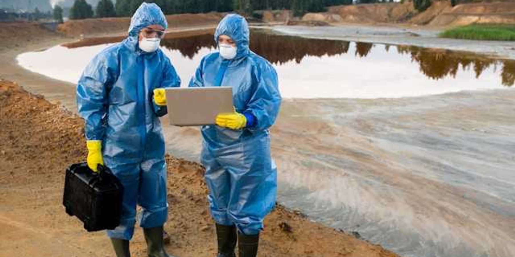 Two people wearing protective gear looking at a laptop in front of a mining waste pond