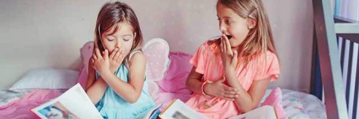 Two small girls sitting on a bed laughing with open books on their laps