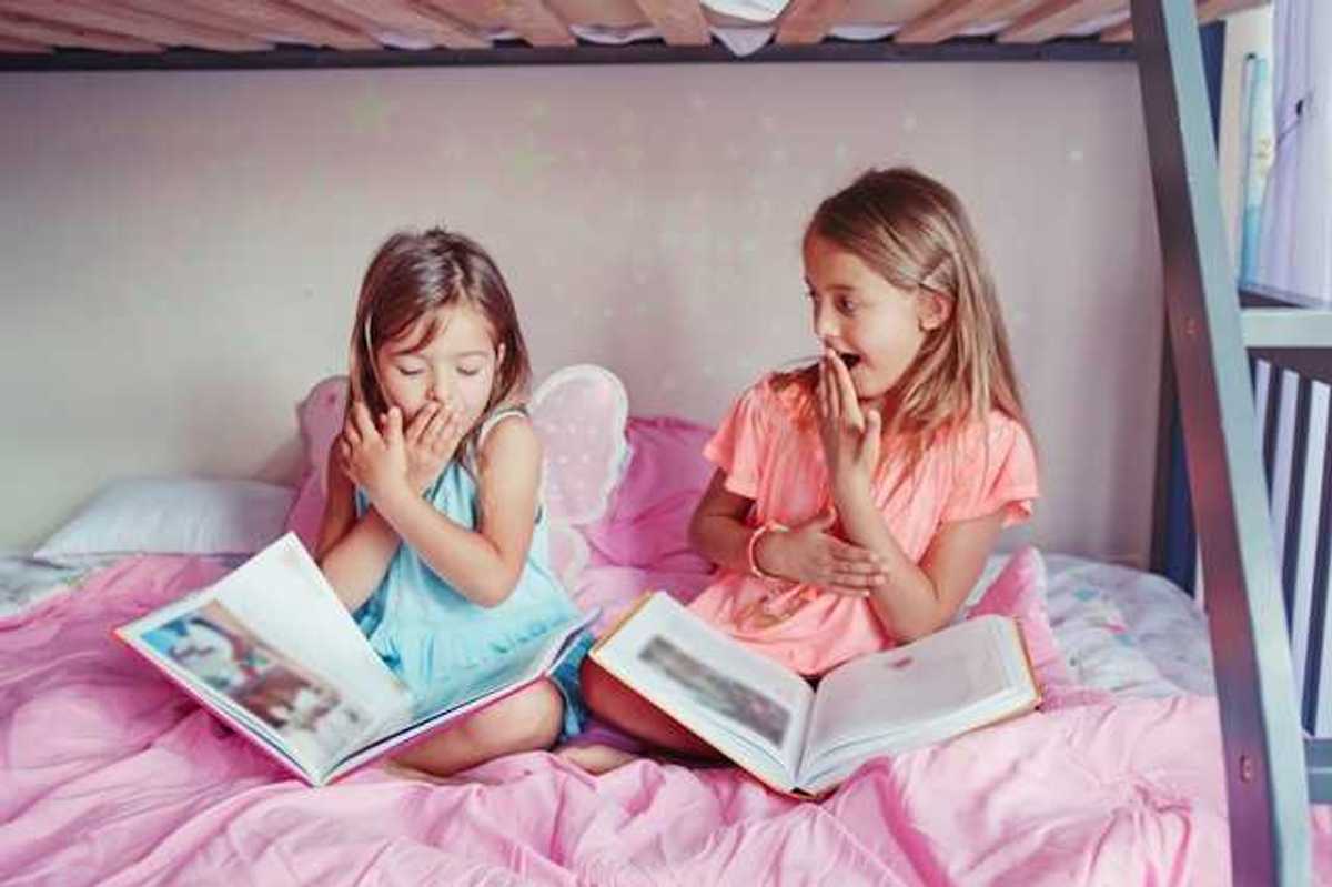 Two small girls sitting on a bed laughing with open books on their laps