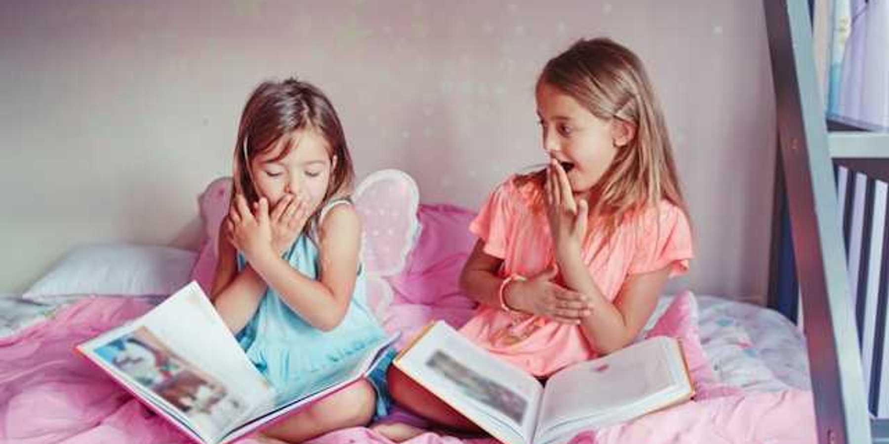 Two small girls sitting on a bed laughing with open books on their laps