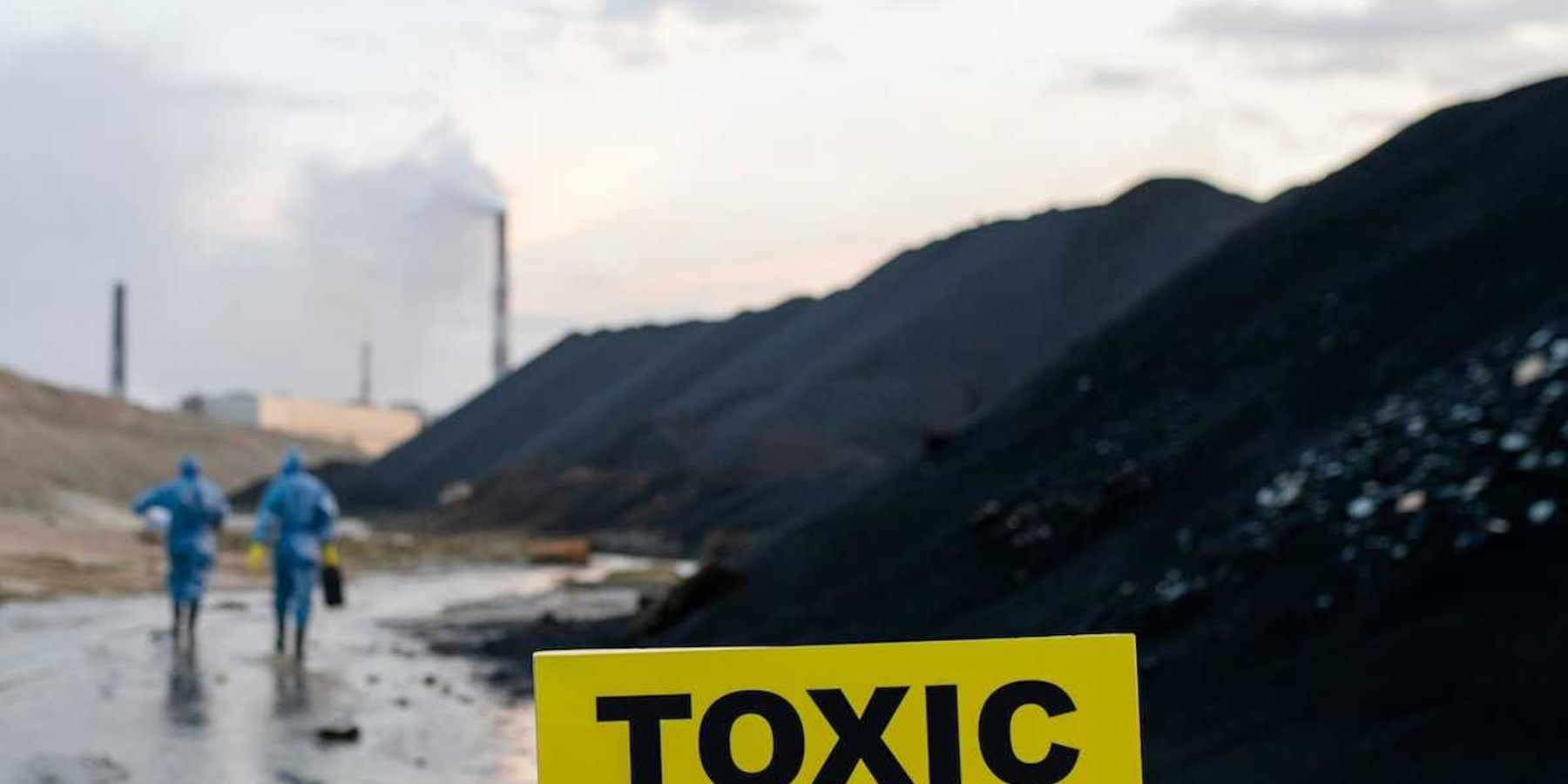 Two workers in blue hazmat suits walking towards coal-fired power plant with sign in foreground warning "toxic danger."