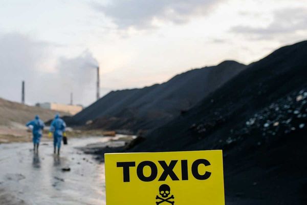 Two workers in blue hazmat suits walking towards coal-fired power plant with sign in foreground warning "toxic danger."