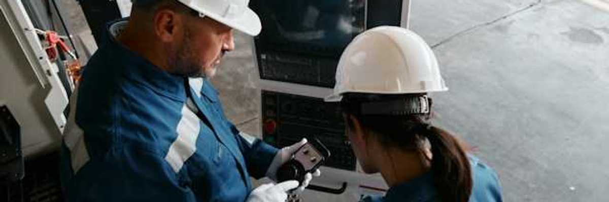Two workers in blue jumpsuits and white hard hats looking at industrial equipment