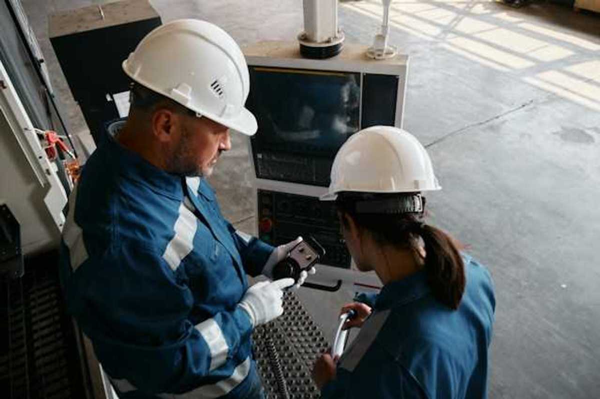 Two workers in blue jumpsuits and white hard hats looking at industrial equipment