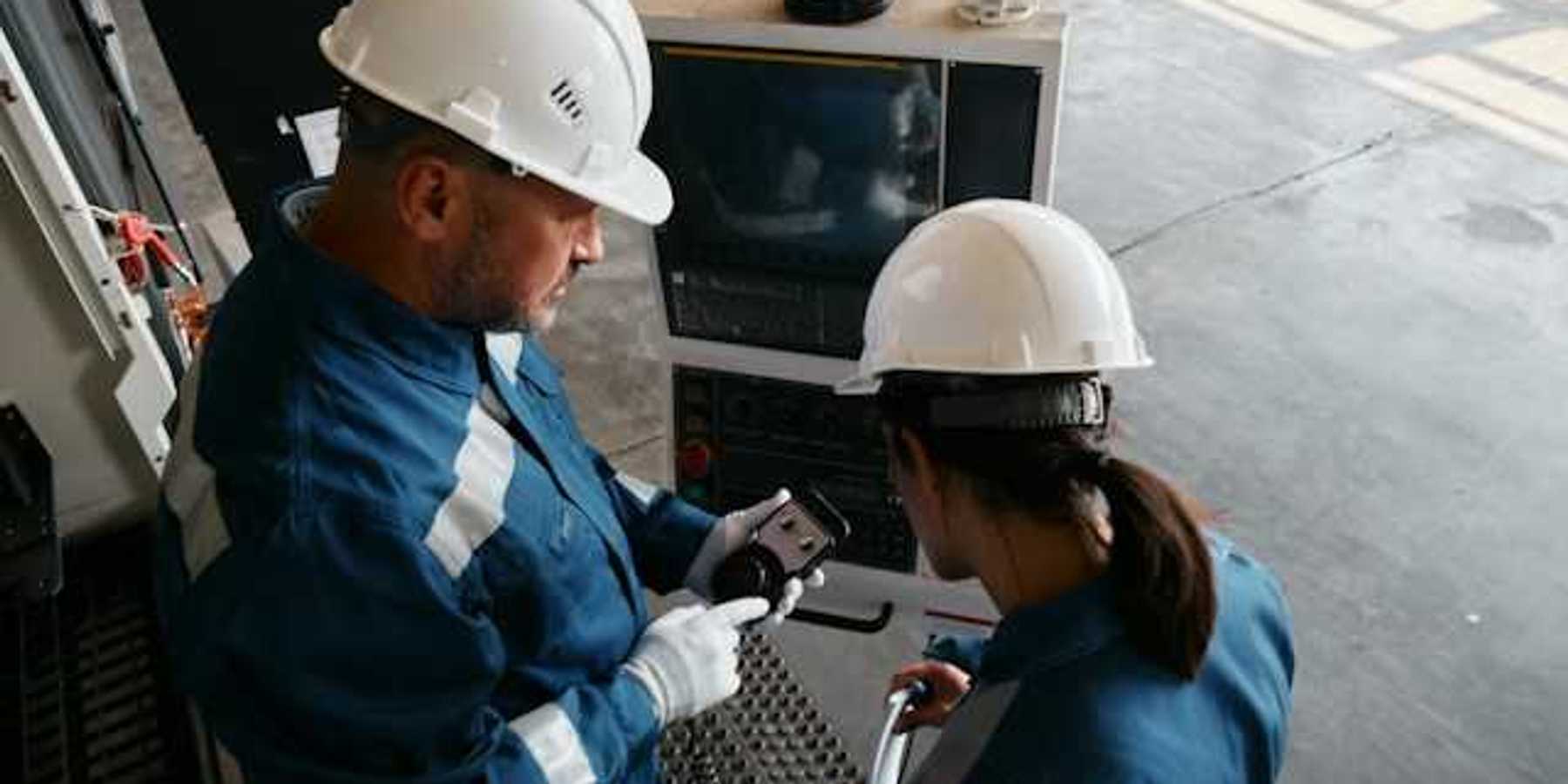Two workers in blue jumpsuits and white hard hats looking at industrial equipment