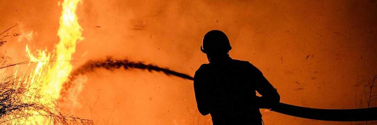 Wildland fire fighter, silhouetted against flame-orange backdrop, training hose on wildfire.