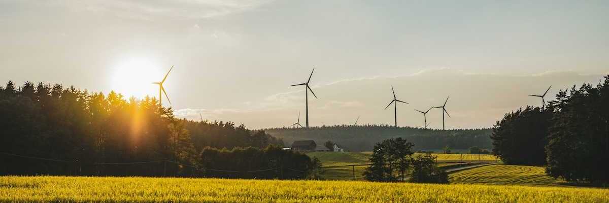Wind turbines sited in a rural setting of forested hills with a farm and meadows in foreground
