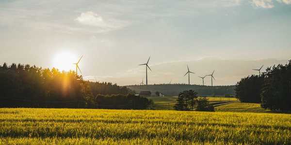 Wind turbines sited in a rural setting of forested hills with a farm and meadows in foreground