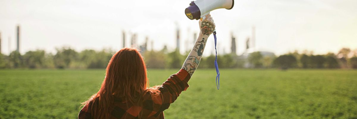 Woman holding up a megaphone facing a refinery across a green field.