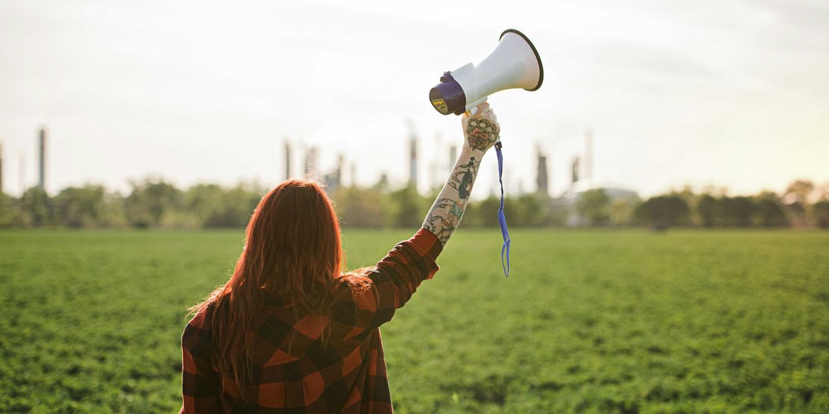 Woman holding up a megaphone facing a refinery across a green field.