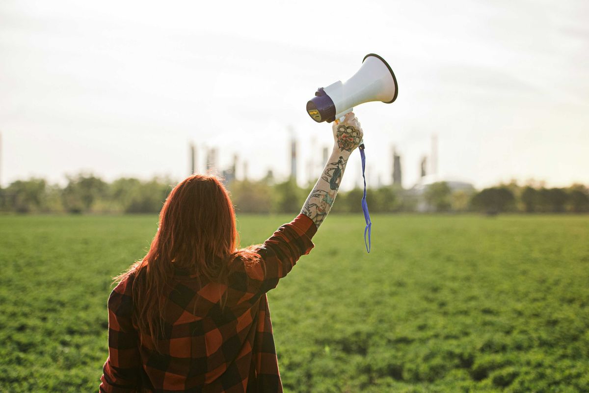 Woman holding up a megaphone facing a refinery across a green field.