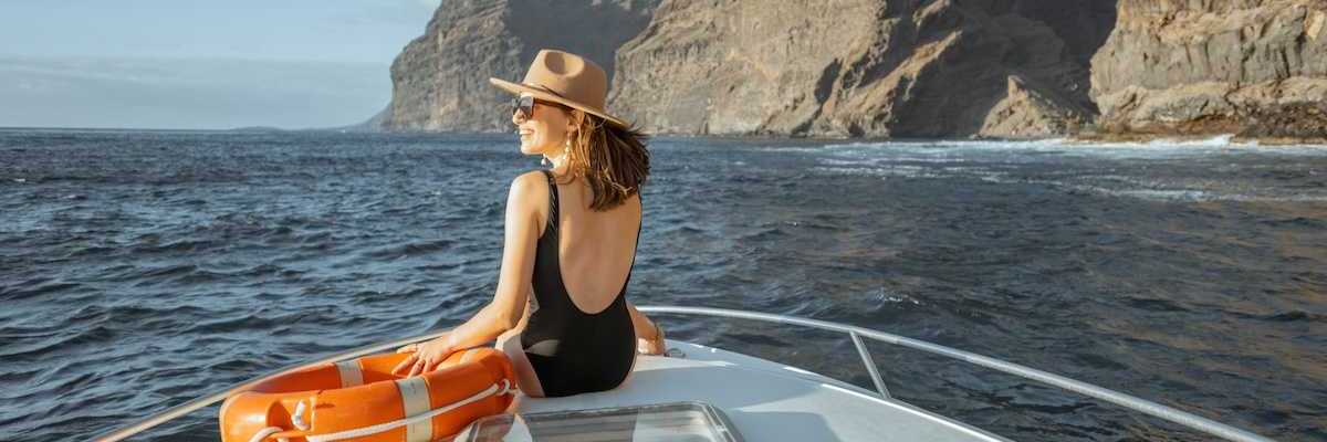 Woman in black bathing suit and broad brimmed hat next to an orange life preserver riding on the bow of a yacht