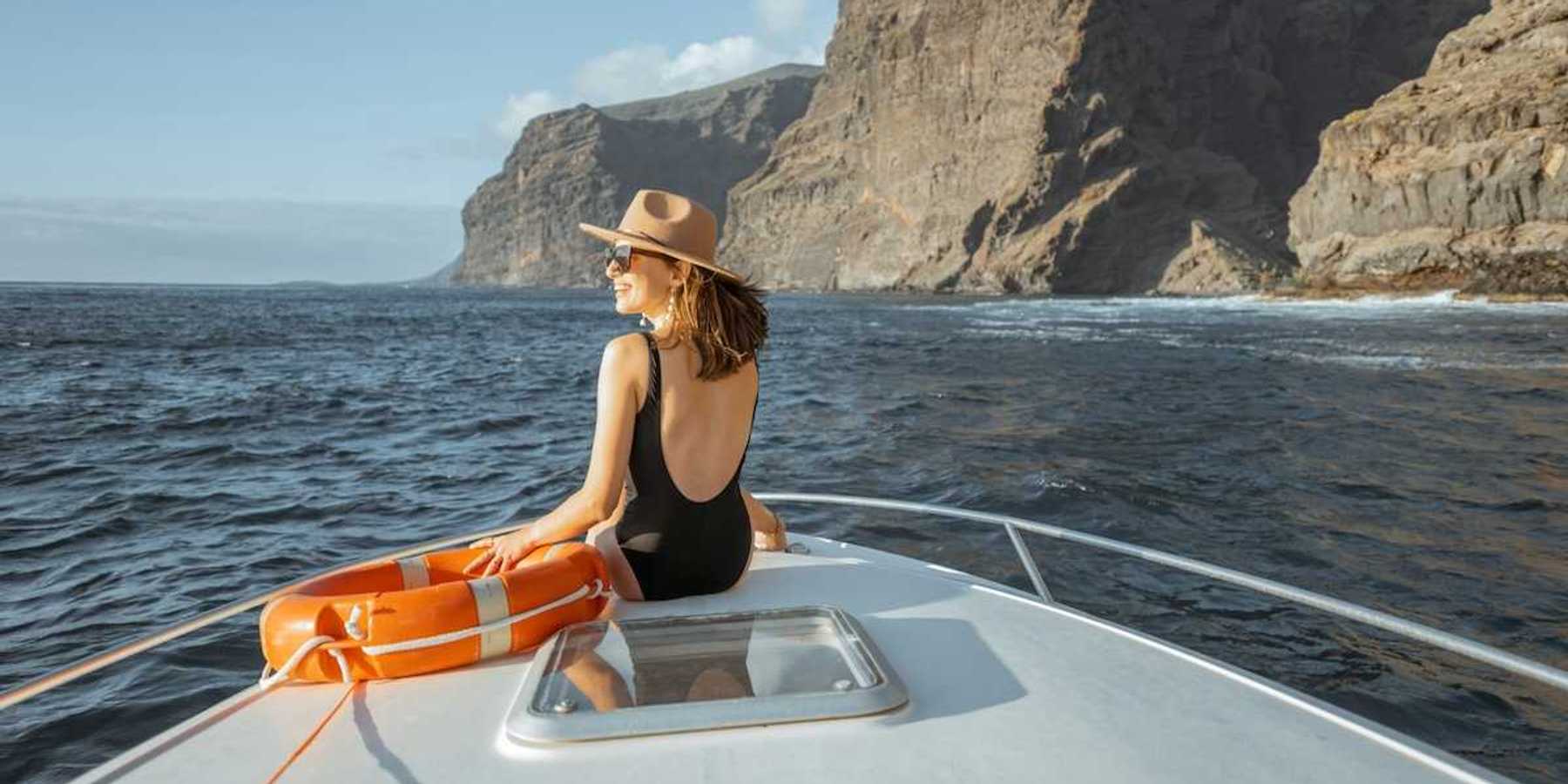 Woman in black bathing suit and broad brimmed hat next to an orange life preserver riding on the bow of a yacht