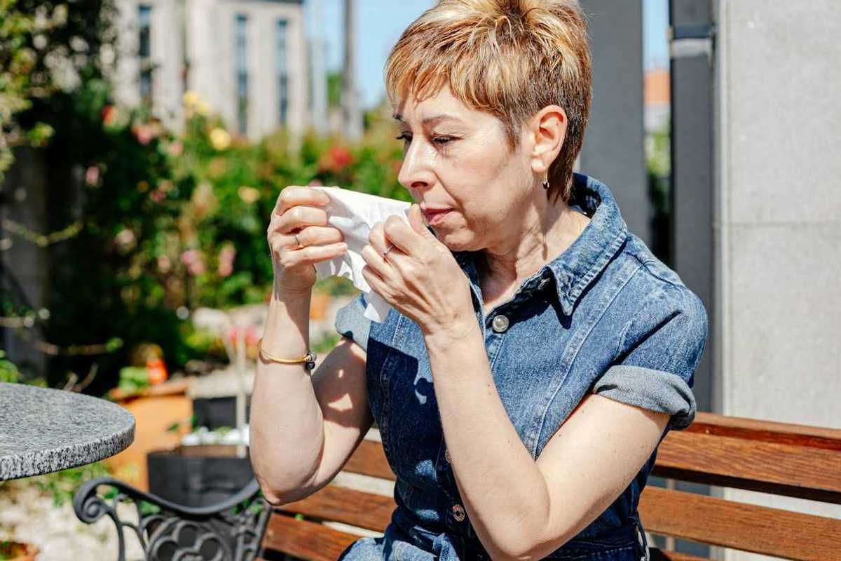 Woman in blue denim dress seated on an outdoor bench preparing to wipe nose with tissue
