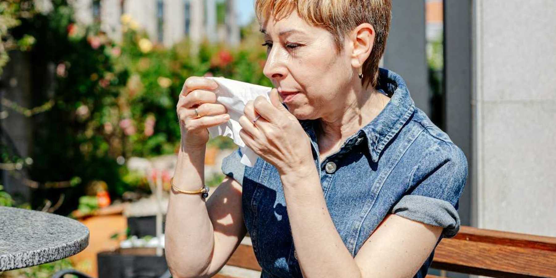 Woman in blue denim dress seated on an outdoor bench preparing to wipe nose with tissue