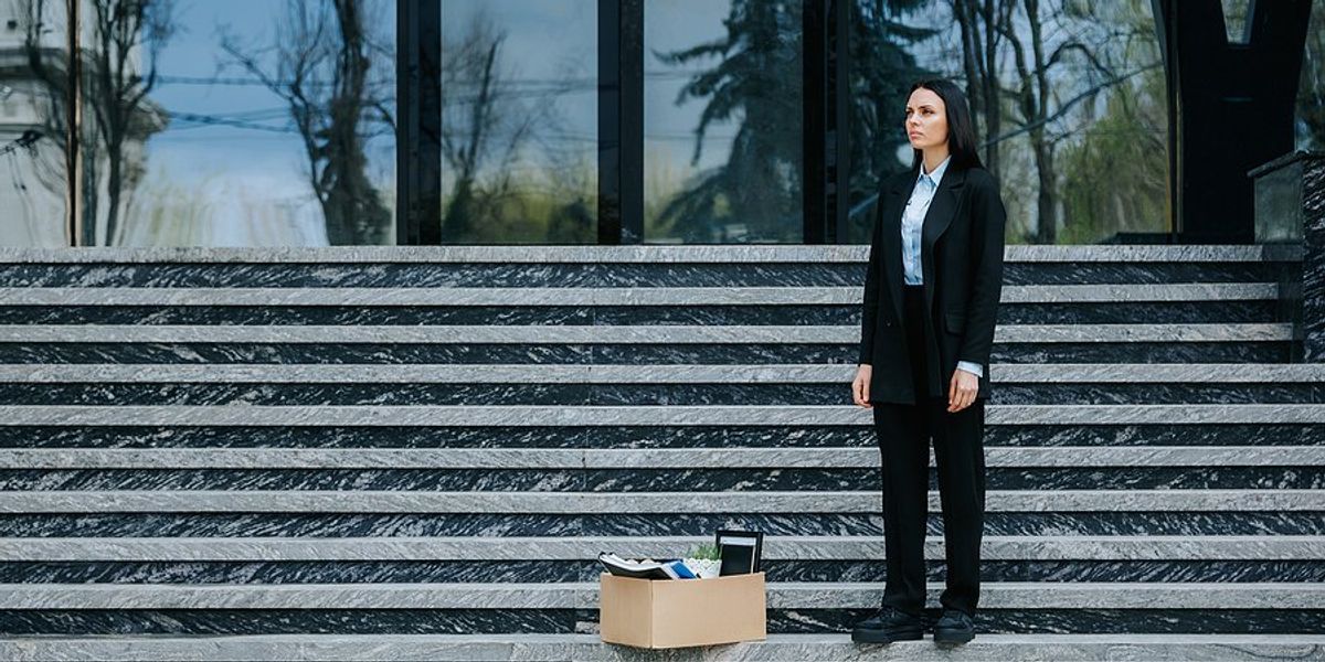 Woman in business attire standing on stairs in front of a building with a box of personal items next to her.