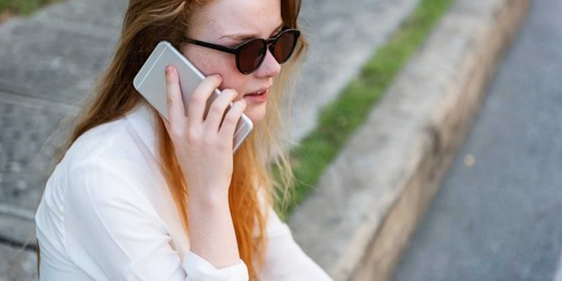 Woman in white shirt and sunglasses sitting on sidewalk listening to something on her phone.