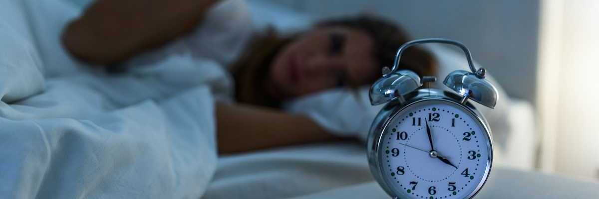Woman lying awake in bed while an alarm clock indicates 3:58, presumably AM, exemplifies poor sleep.