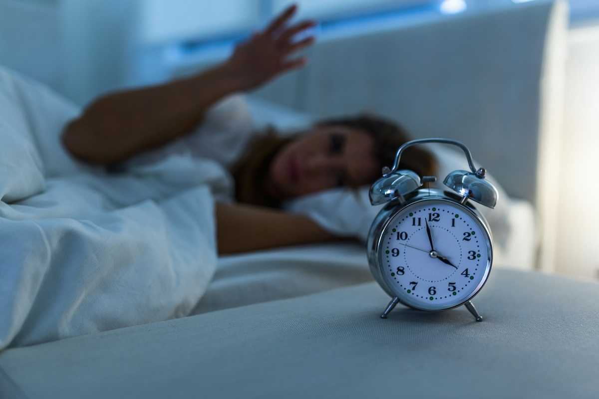 Woman lying awake in bed while an alarm clock indicates 3:58, presumably AM, exemplifies poor sleep.