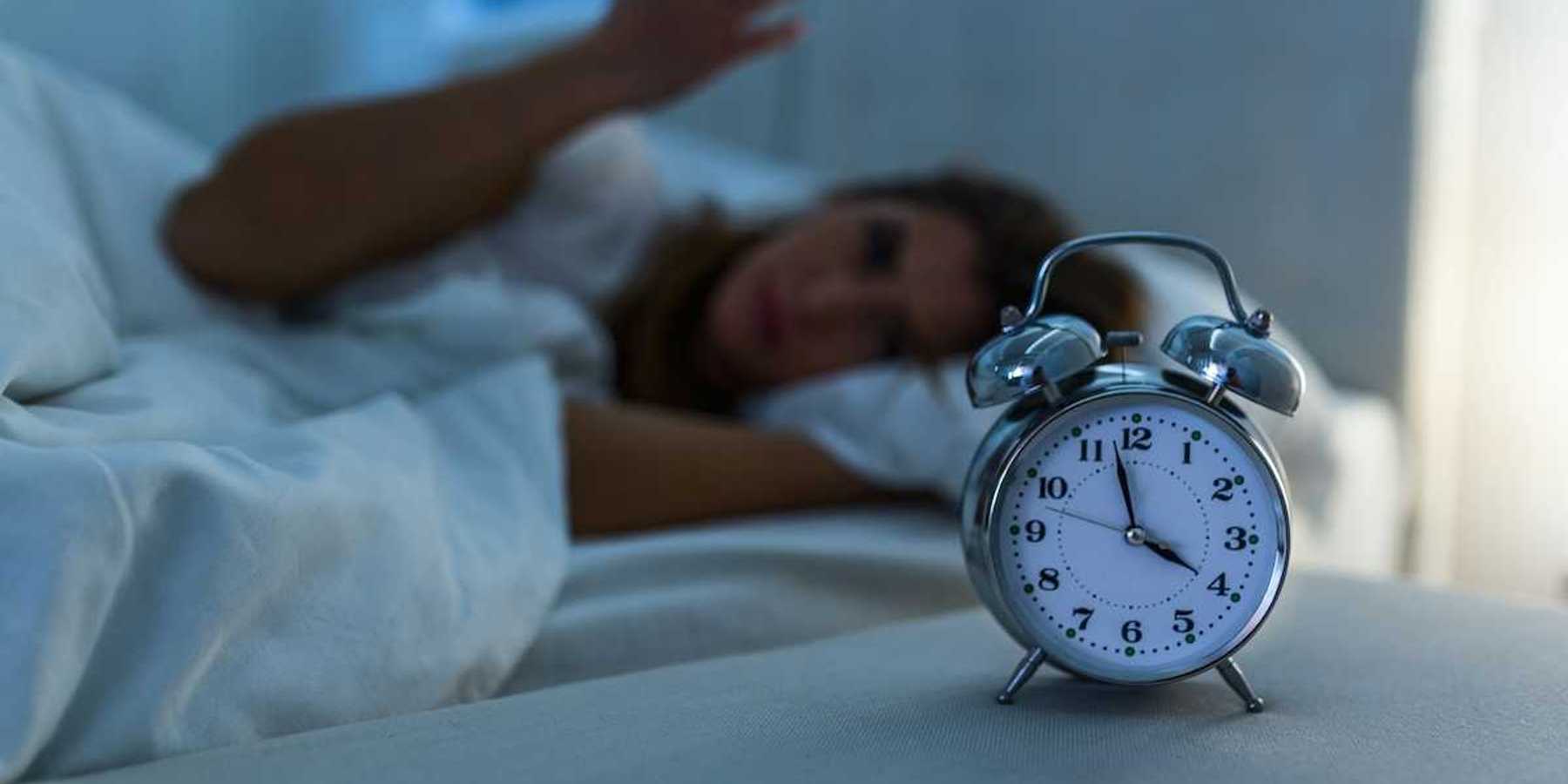 Woman lying awake in bed while an alarm clock indicates 3:58, presumably AM, exemplifies poor sleep.