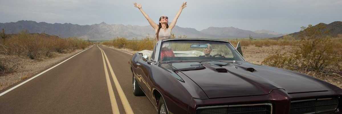 Woman riding in "muscle car" convertible, either standing or sitting on seat rest with arms upraised implying freedom of the road