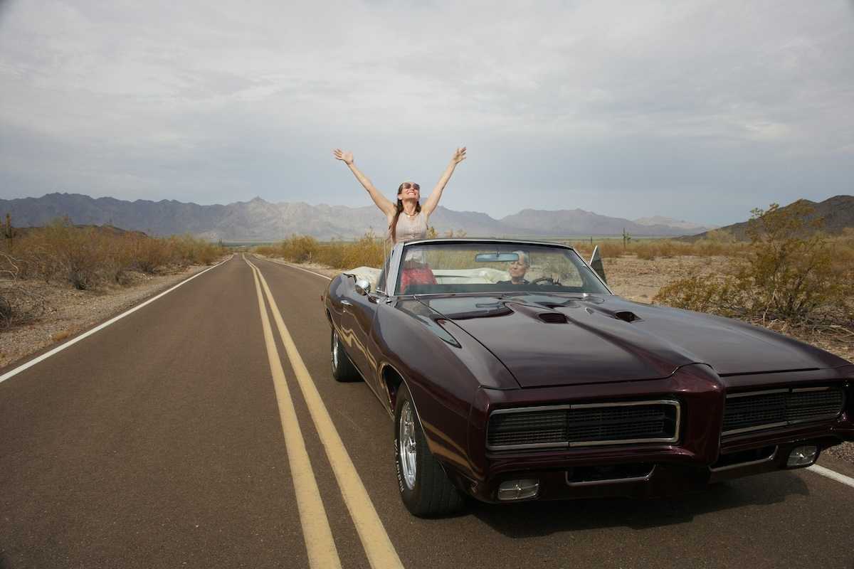 Woman riding in "muscle car" convertible, either standing or sitting on seat rest with arms upraised implying freedom of the road