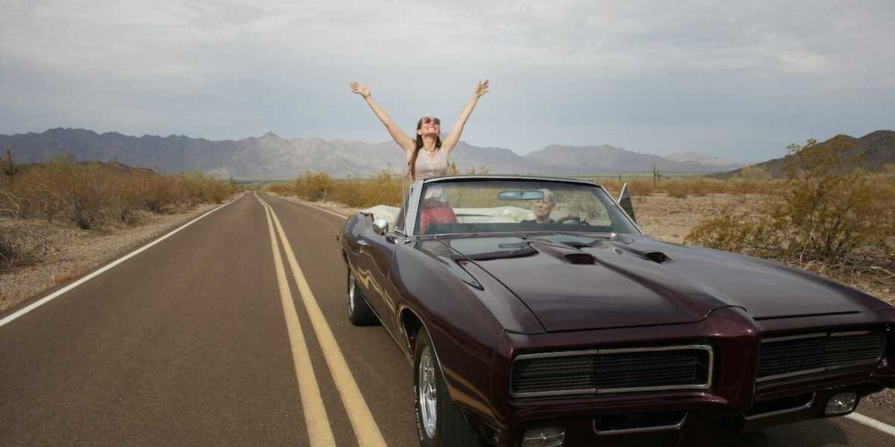 Woman riding in "muscle car" convertible, either standing or sitting on seat rest with arms upraised implying freedom of the road