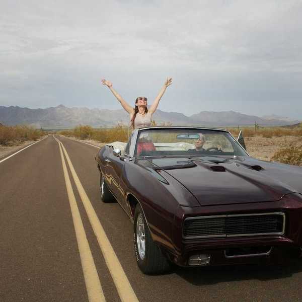 Woman riding in "muscle car" convertible, either standing or sitting on seat rest with arms upraised implying freedom of the road