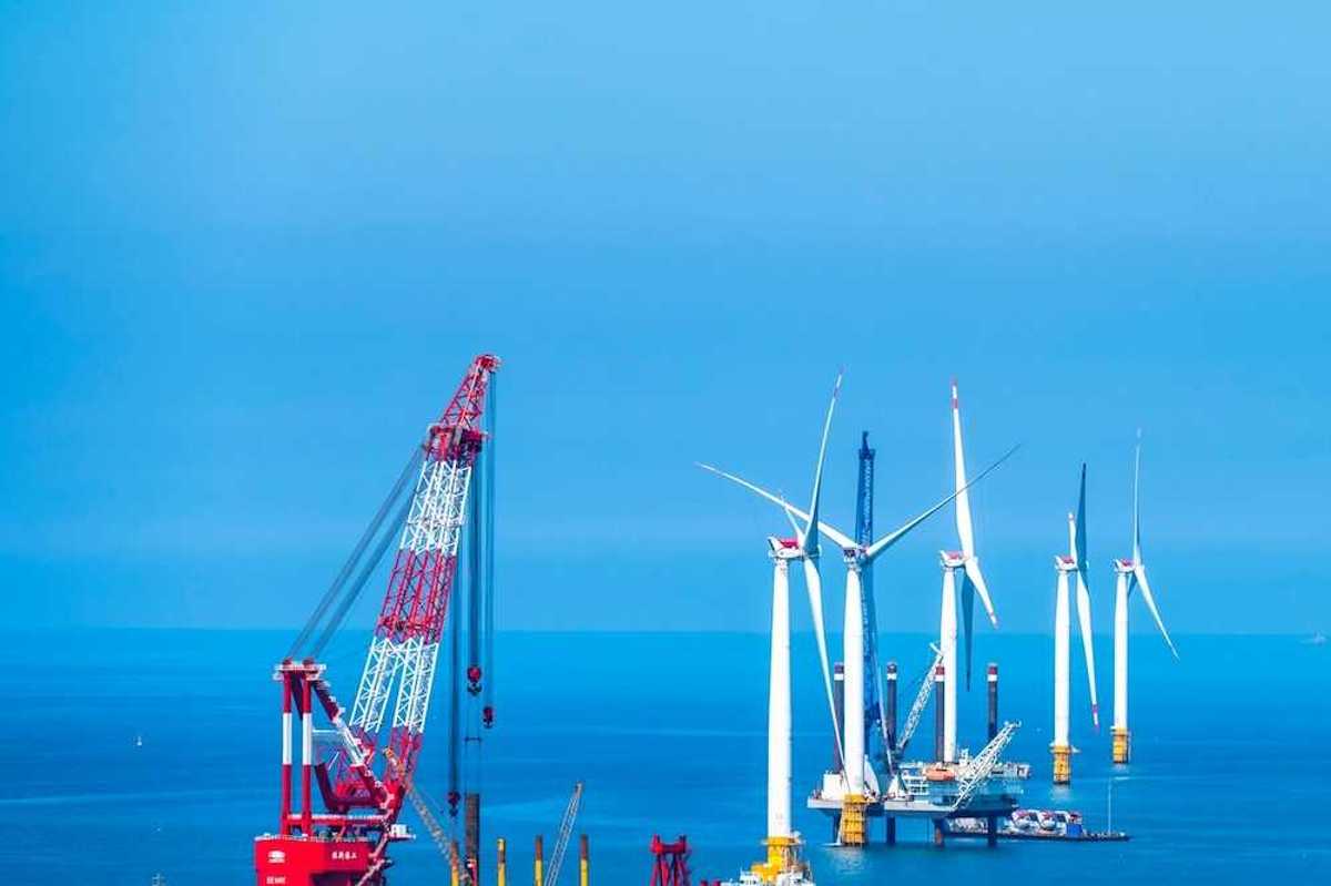 Yellow and white offshore wind turbines being installed using a floating red & white crane
