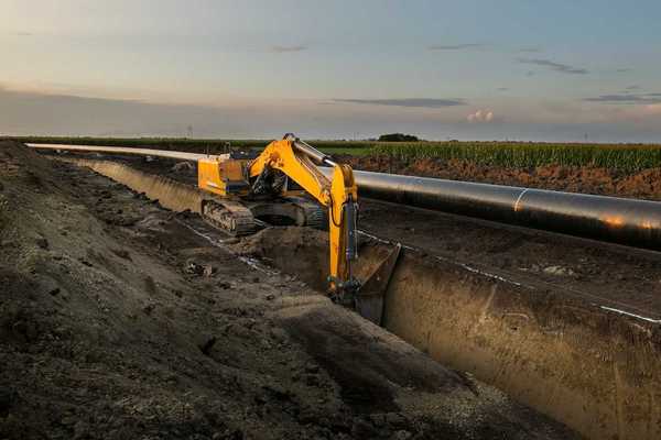 Yellow excavator digging deep trench making way for pipeline construction