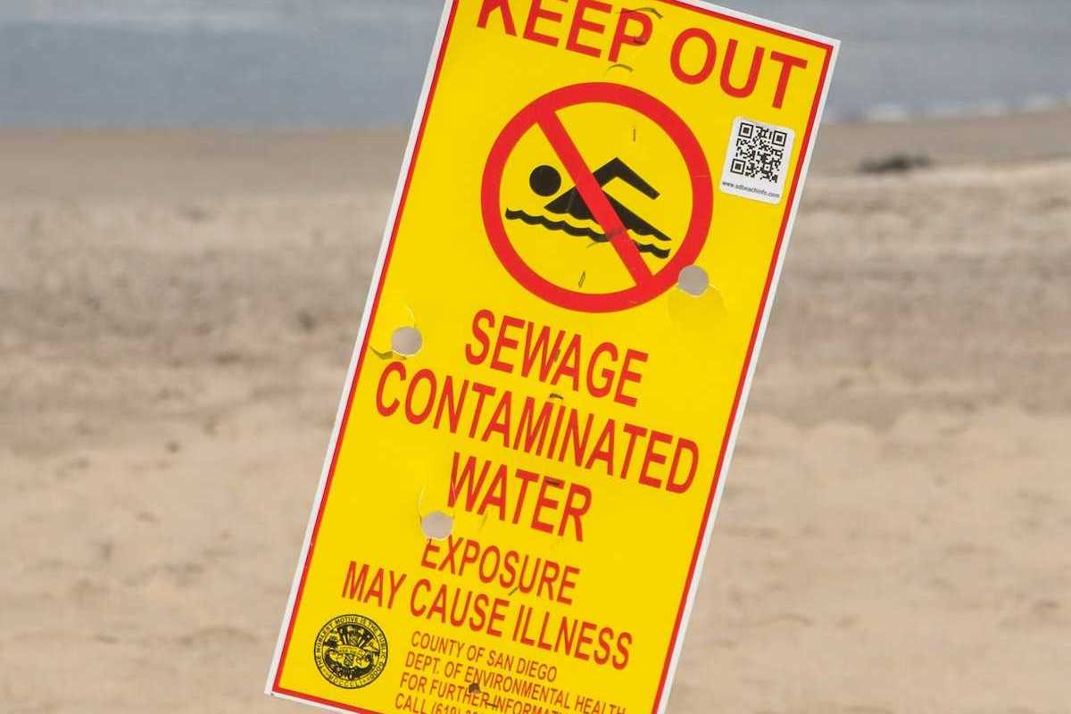 Yellow sign with red lettering reads "KEEP OUT, SEWAGE CONTAMINATED WATER. EXPOSURE MAY CAUSE ILLNESS"