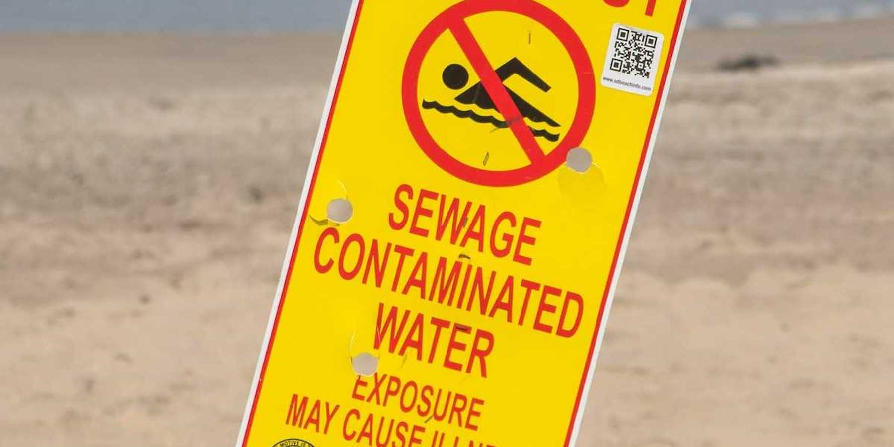 Yellow sign with red lettering reads "KEEP OUT, SEWAGE CONTAMINATED WATER. EXPOSURE MAY CAUSE ILLNESS"