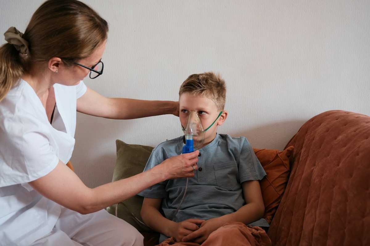 Young boy seated on sofa fitted with an inhaler being adfministered by a nurse in white