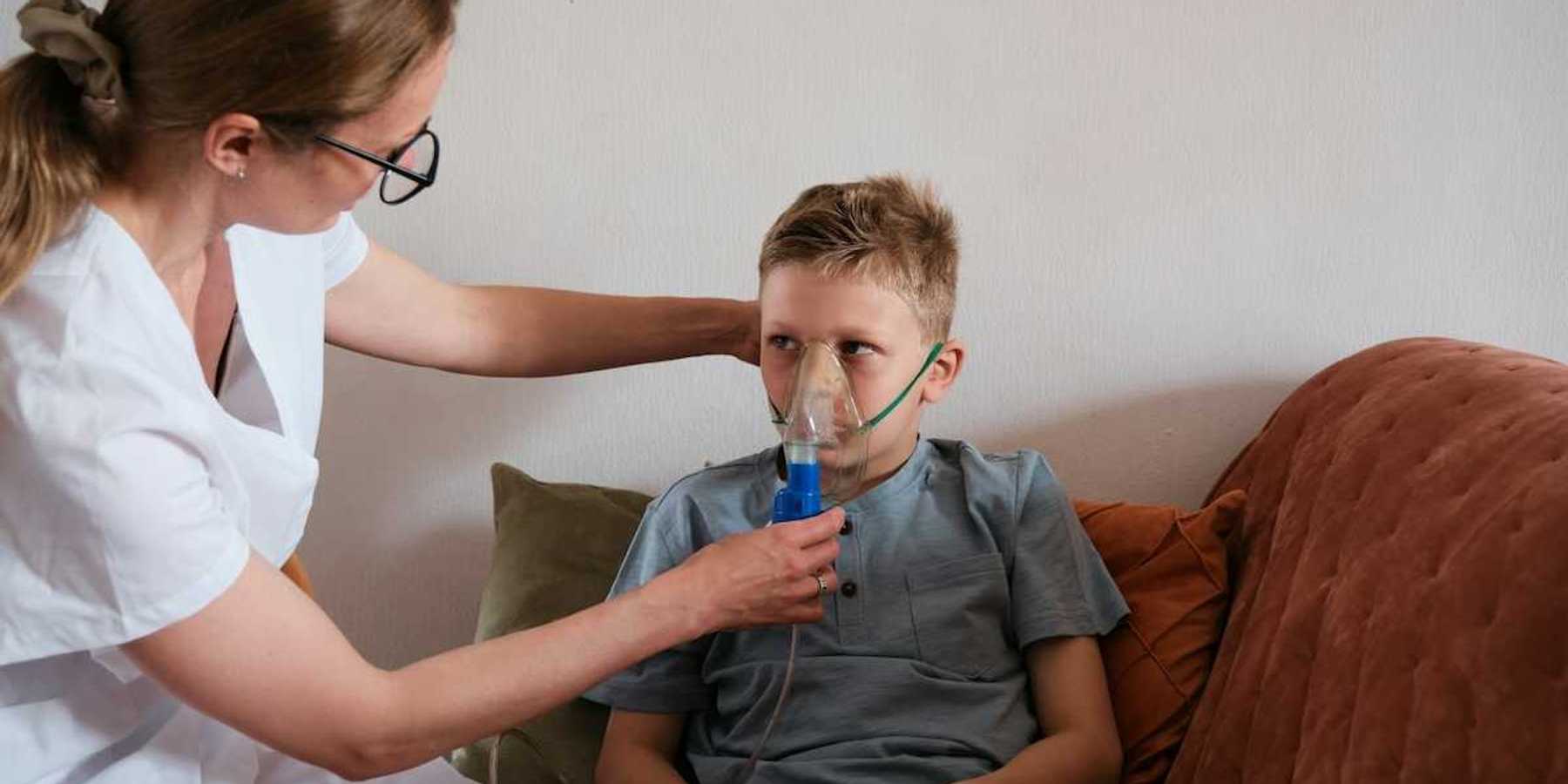 Young boy seated on sofa fitted with an inhaler being adfministered by a nurse in white