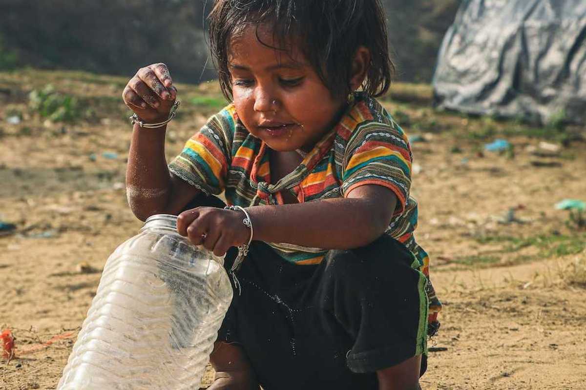 Young child squatting in the dirt, adding coins to a plastic jar