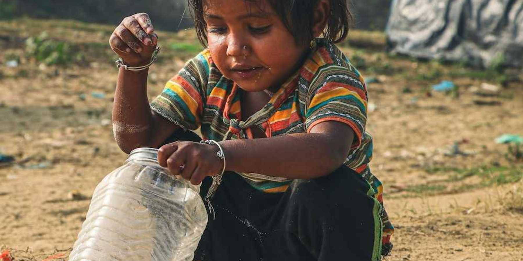 Young child squatting in the dirt, adding coins to a plastic jar